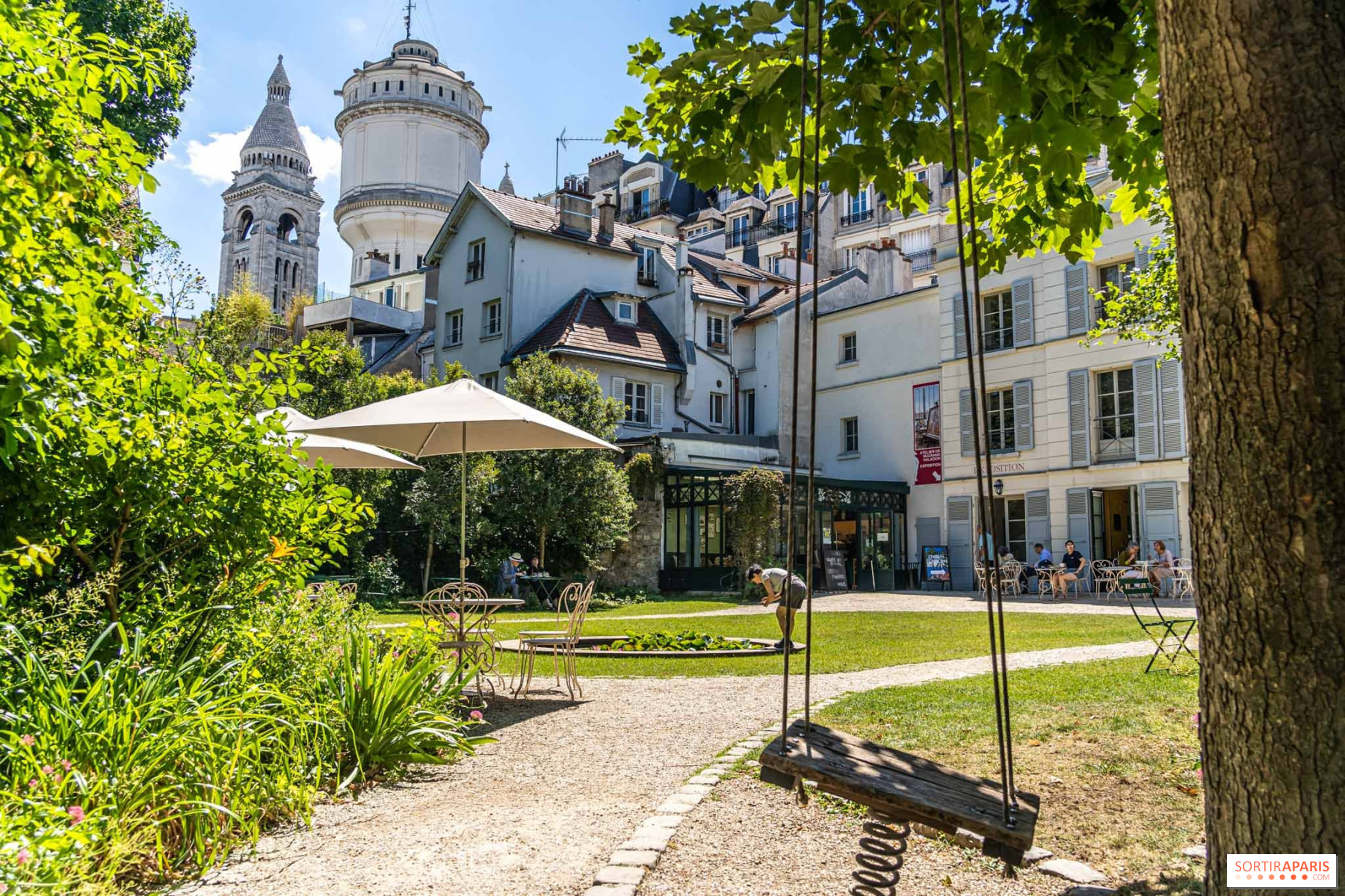 A beautiful Parisian garden at the Musée de Montmartre featuring a rustic wooden swing in the foreground and historic buildings in the background under a clear sky.